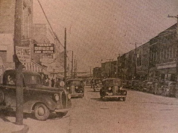 Sepia photograph of West Main Street with early automobiles parked along the storefronts.