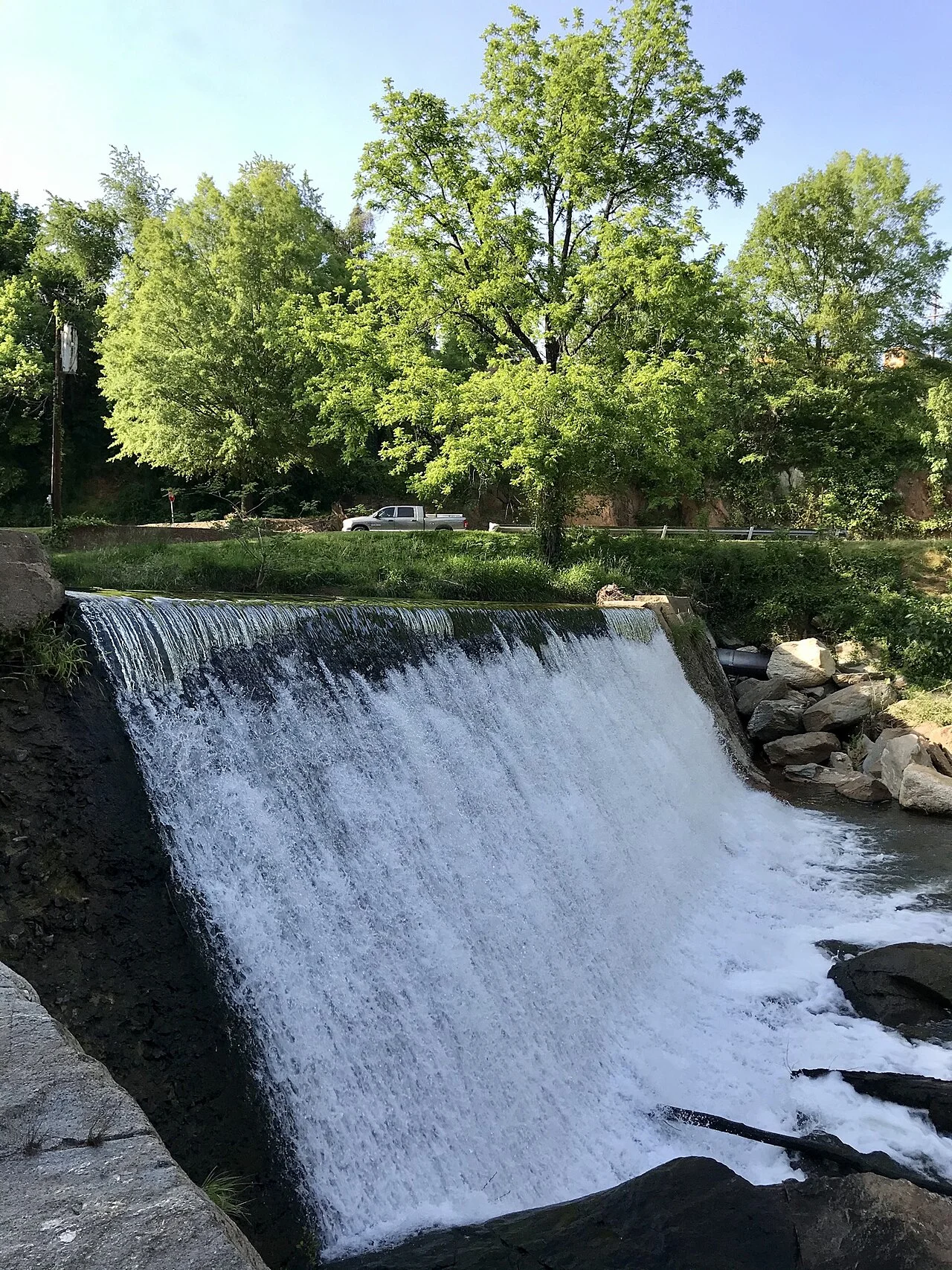 Water pours over an old stone mill dam among leafy trees.