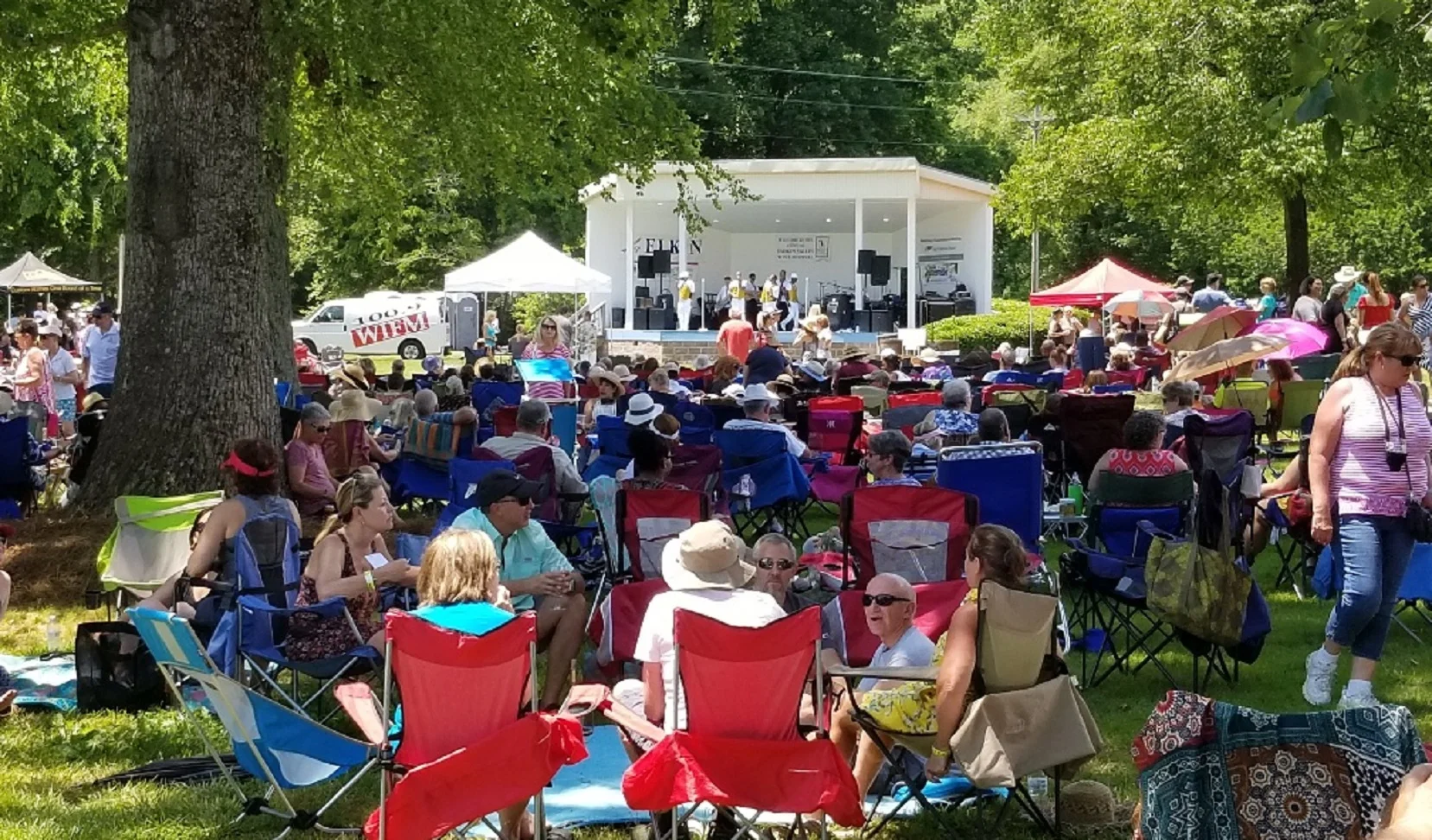A festival crowd in lawn chairs faces a white band shell where a band plays under tall trees.