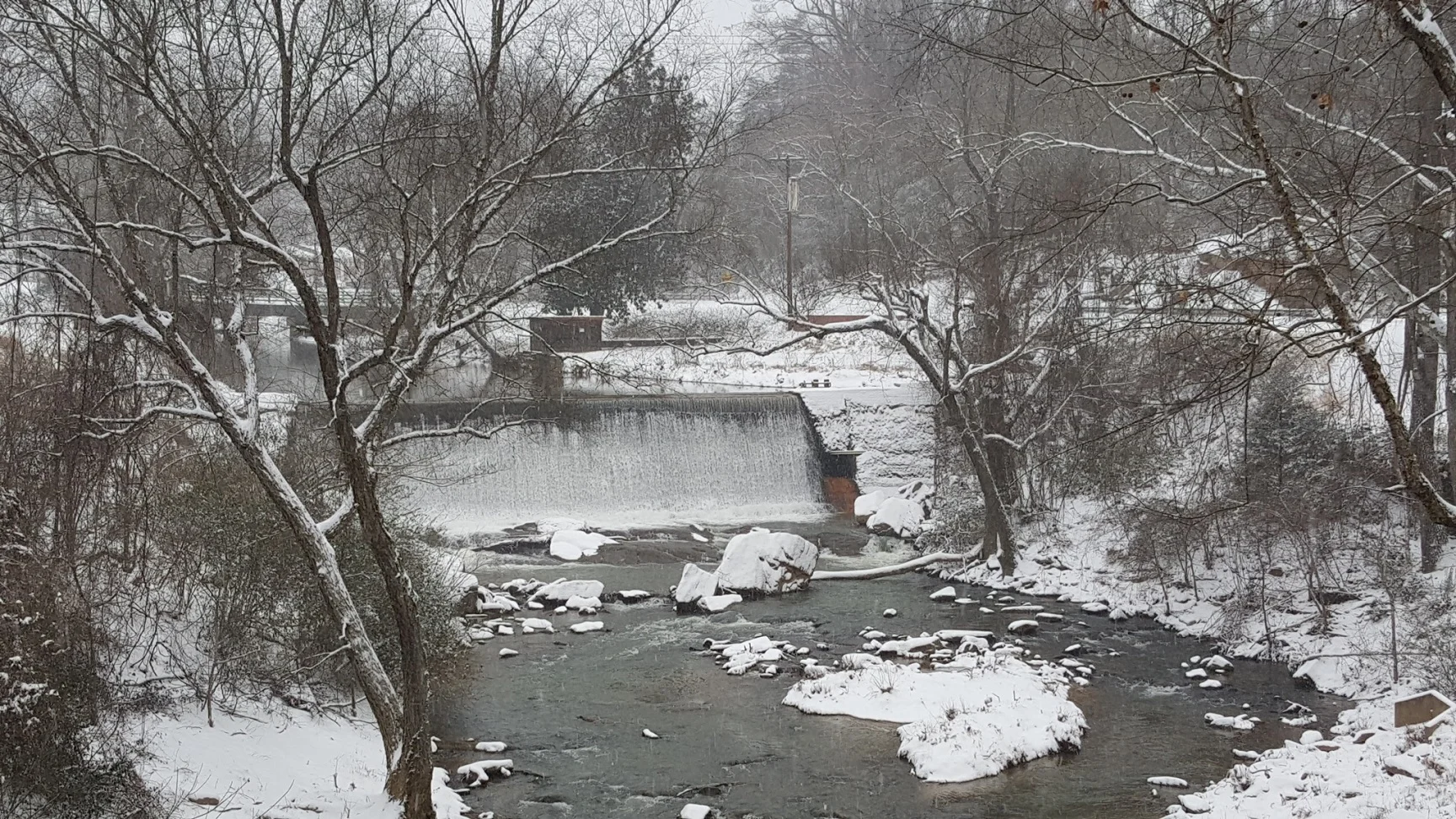 Snow covers the rocks and banks of a creek below a wide stone dam, with bare trees along the water.