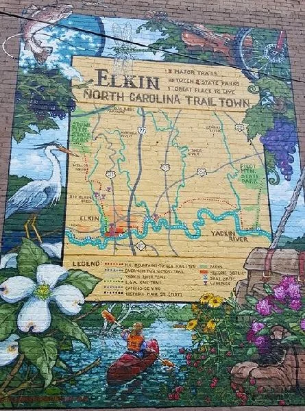 A mural painted on brick: a sign reading "Elkin, North Carolina Trail Town" with the lines "3 major trails, between 2 state parks, 1 great place to live", framed by flowers, a dragonfly, and a fiddle.