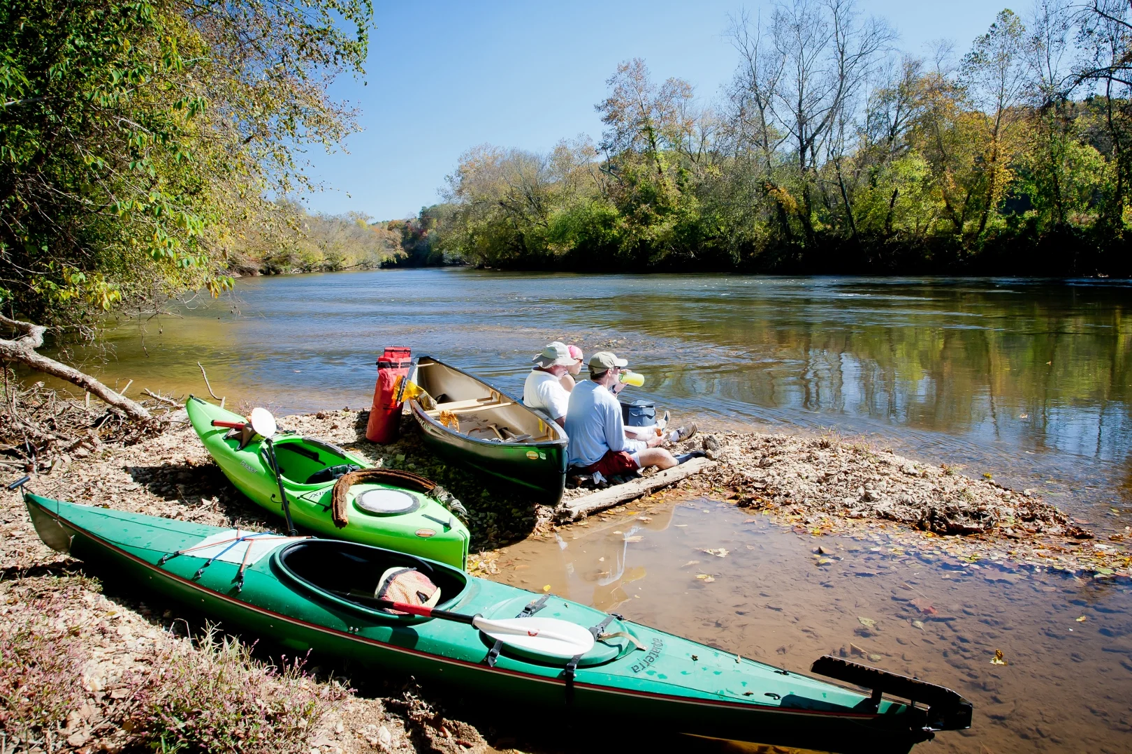 Paddlers beside their kayaks on the Yadkin River, with autumn trees across the water.