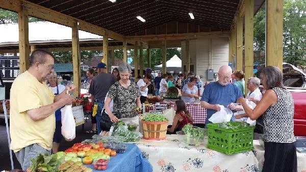 Shoppers and vendors crowd tables of produce under the open-sided Farmers Market shelter.