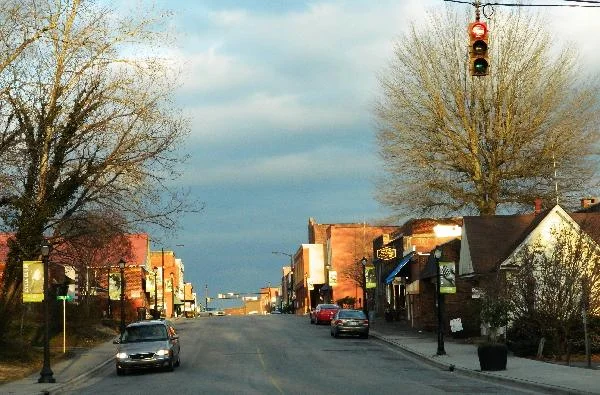 Brick storefronts line Main Street in spring, under early leaves and a traffic signal.