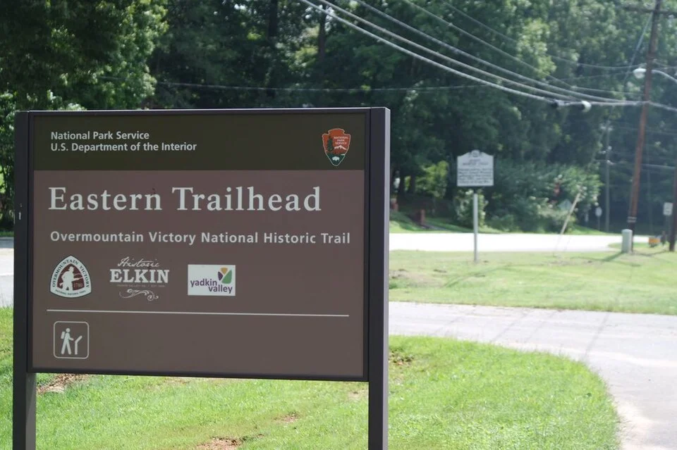 A National Park Service sign marks the Eastern Trailhead of the Overmountain Victory National Historic Trail beside a paved path.