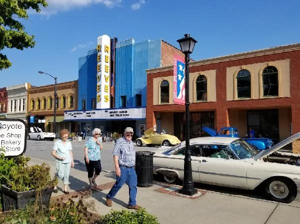 People walk along West Main Street past brick storefronts, the Reeves Theater sign, and classic cars at the curb.