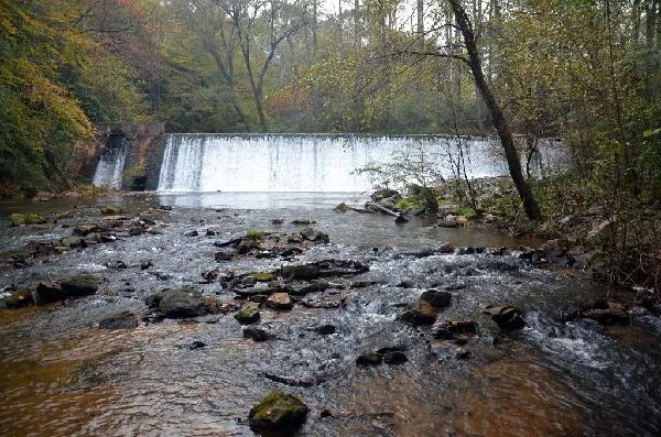 Water spills over the wide, low Shoe Factory Dam into a rocky creek bed in autumn woods.